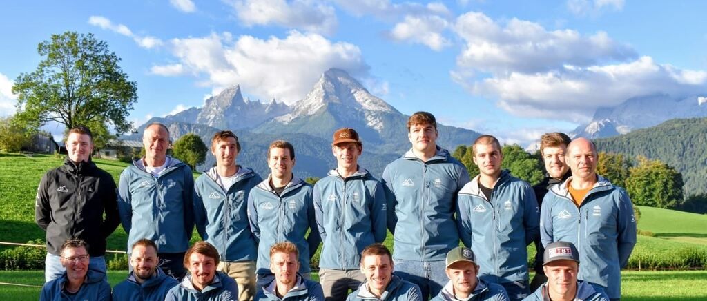A group of men in matching blue jackets and caps pose for a photo in front of a mountainous landscape.