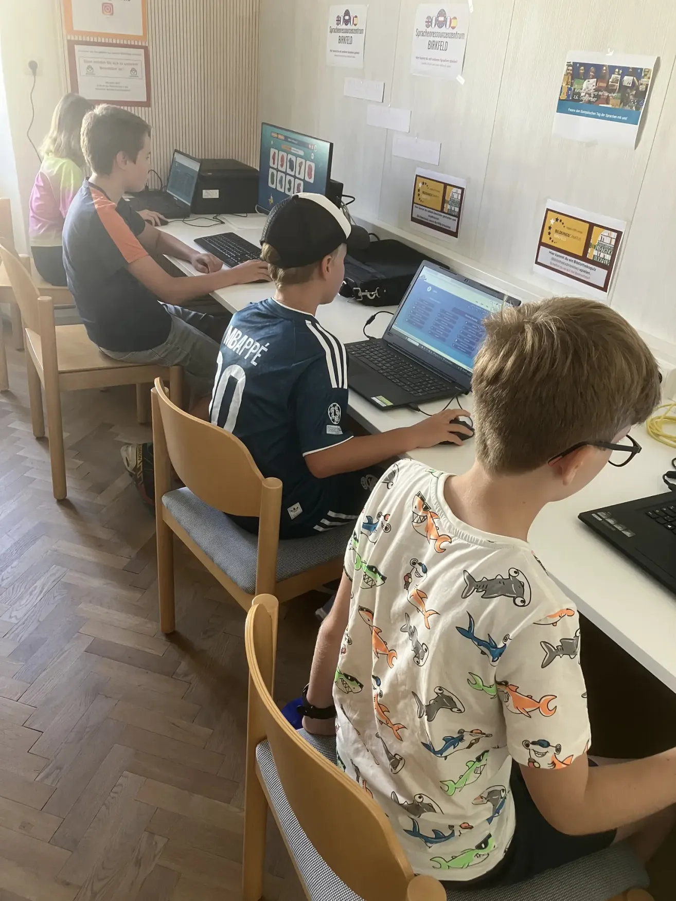 Four boys sit at computers in a classroom, each focused on their screens. The room has wooden floors and white walls.