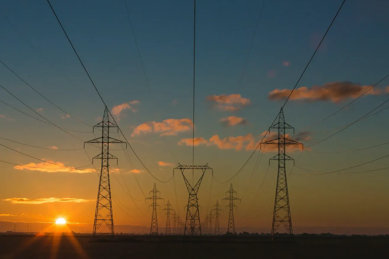 Eine Reihe hoher elektrischer Übertragungsmasten vor einem Himmel mit warmen Farbtönen bei Sonnenuntergang.