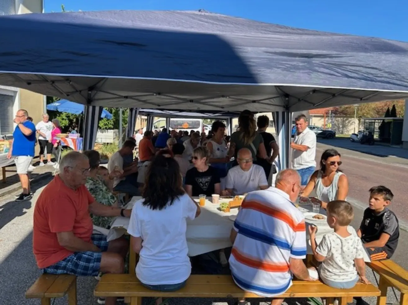 Eine Gruppe von Menschen versammelt sich unter einem Zelt für eine Mahlzeit, einige sitzen auf Bänken und andere stehen. Der Tisch ist mit Tellern, Tassen und Essen gedeckt. Die Umgebung ist im Freien, mit einem klaren blauen Himmel und Bäumen im Hintergrund.