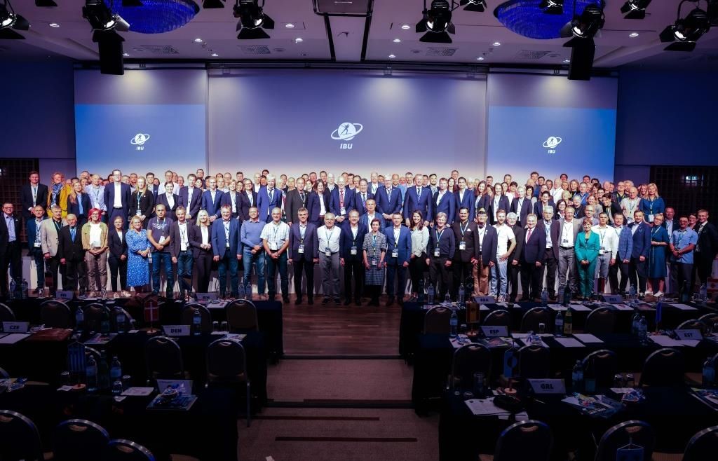 A large group of formally dressed individuals stand in rows in front of a stage with large screens behind them. The screens display logos and text, and the setting appears to be a conference or meeting.
