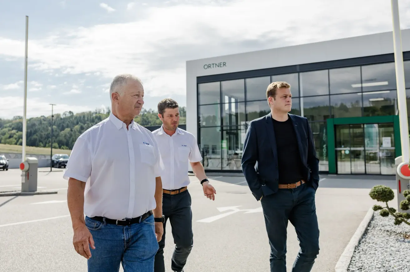 Three men walk in front of a modern building with glass walls, one wearing a white shirt and jeans, another in a white shirt and black pants, and the third in a dark suit.