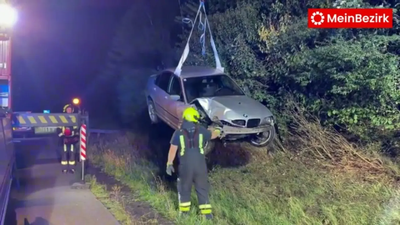 A person in a yellow helmet is pulling a damaged car from the grass. The car is hanging from a rope.
