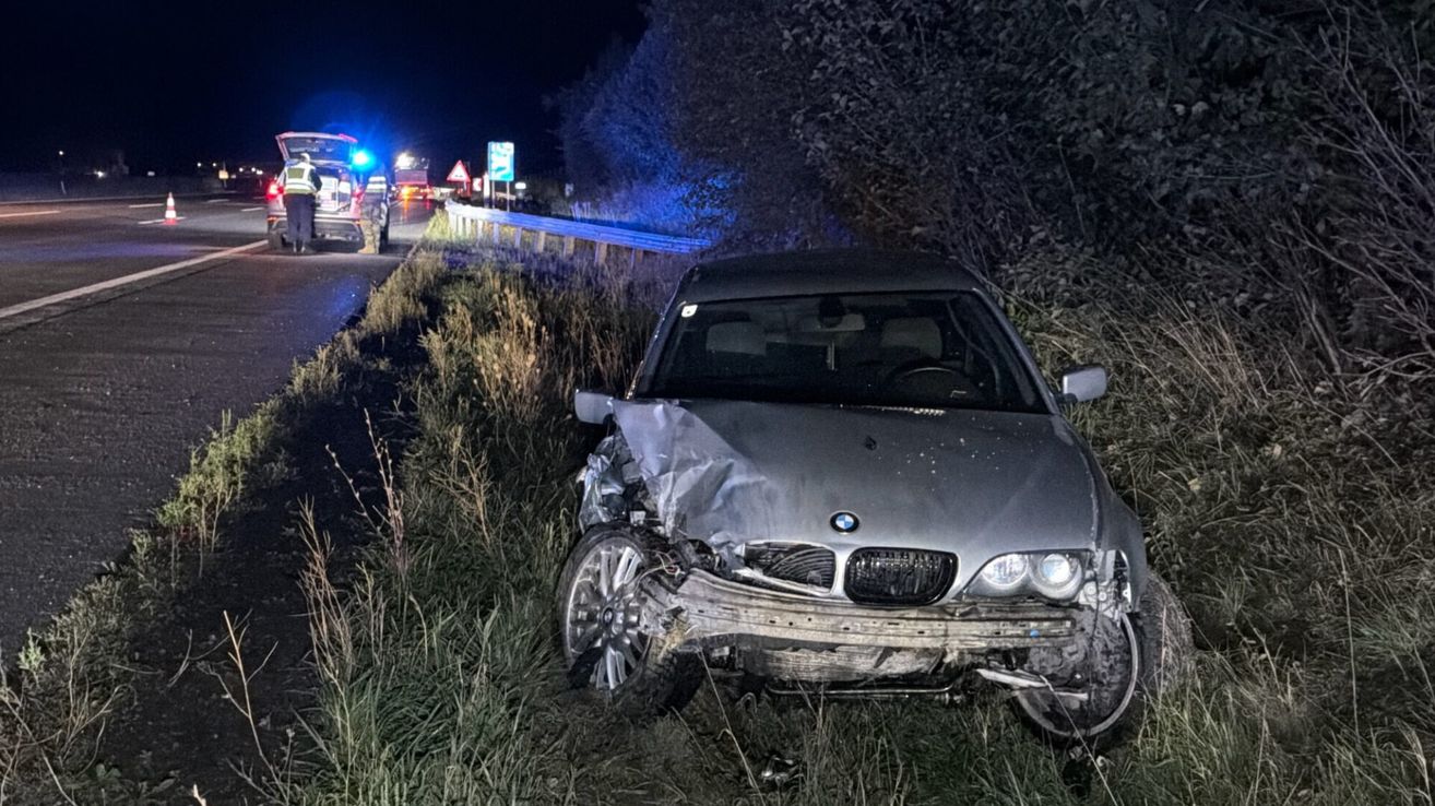A damaged BMW car is parked on the side of a road at night. The front is severely damaged, with the bumper and headlights crushed.
