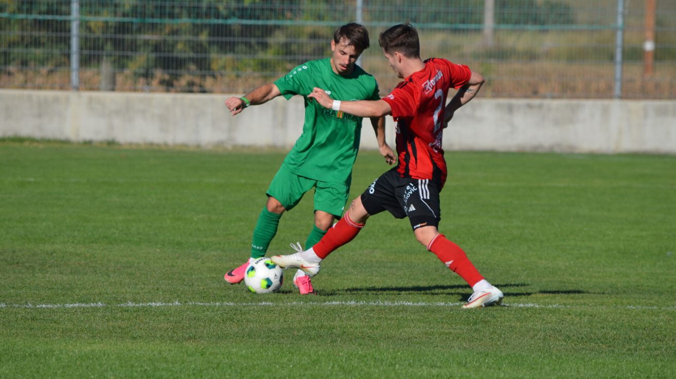 Zwei Fußballspieler, einer in Grün und der andere in Rot, sind in einem Spiel auf einem Rasenfeld engagiert. Der Spieler in Grün versucht, dem Spieler in Rot den Ball zu stehlen.