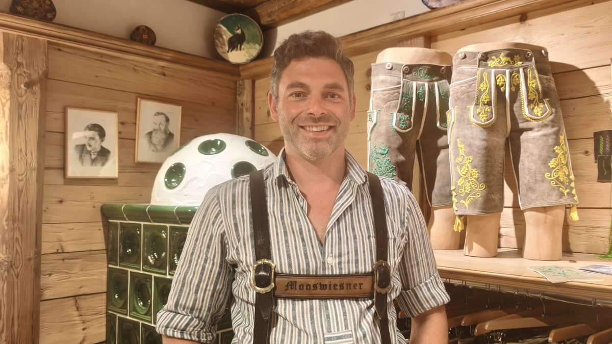 A man in traditional Bavarian attire stands in a room with wooden walls, smiling. Behind him, a large white object and two mannequins wearing embroidered pants are visible.