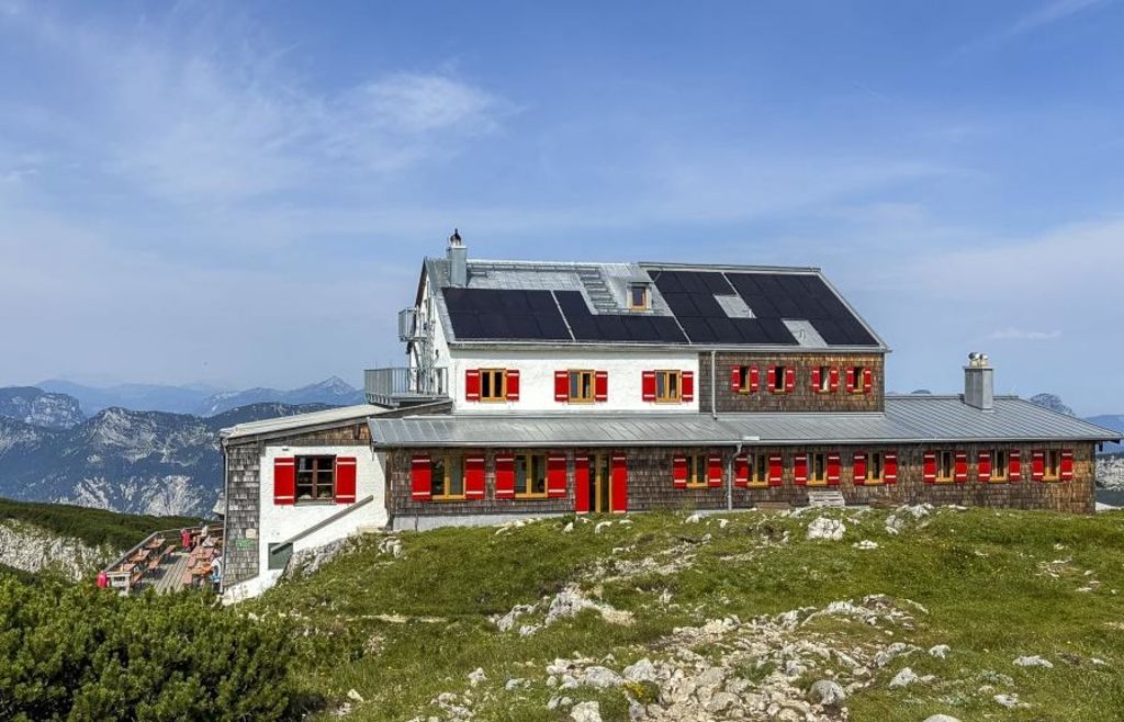 A mountain lodge with solar panels on the roof, red shutters, and a scenic mountain backdrop.