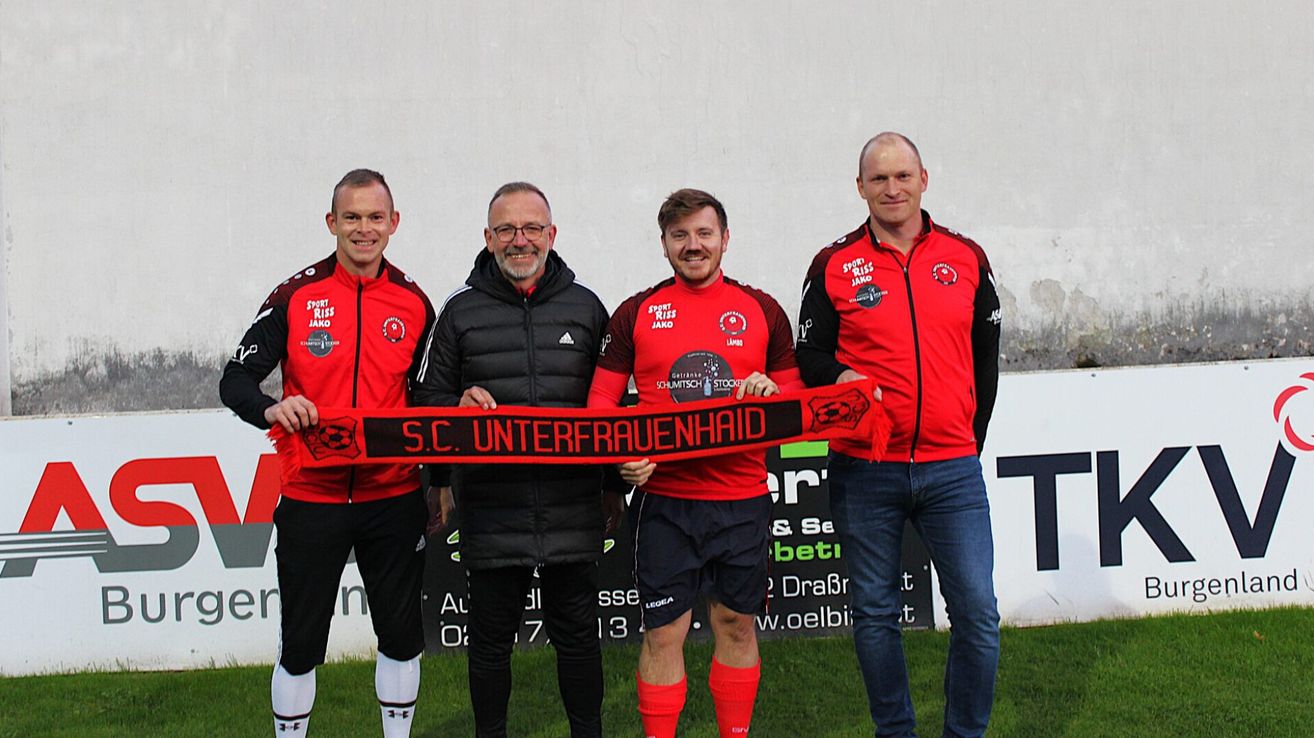 Vier Männer stehen auf einem Rasenfeld und halten ein rotes Banner mit dem Text S.C. Unterfrauenhaid. Sie alle lächeln und scheinen für ein Foto zu posieren.