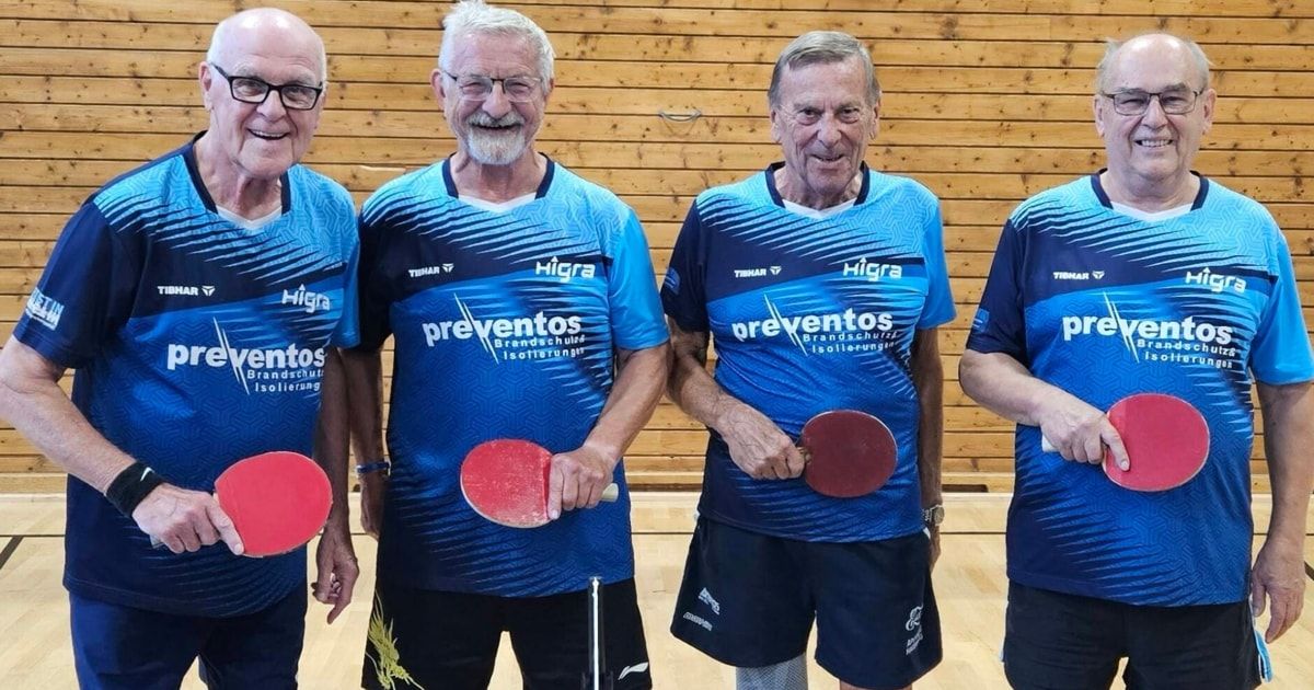 Vier ältere Männer in blauen Sporttrikots mit dem Wort 'Preventos' darauf stehen zusammen und halten Tischtennisschläger. Sie lächeln und scheinen für ein Foto in einem holzverkleideten Raum zu posieren.