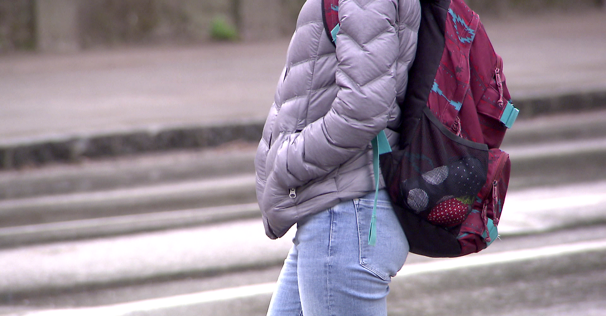 Eine Person in einer grauen Daunenjacke und blauen Jeans geht mit einem roten Rucksack die Straße entlang.