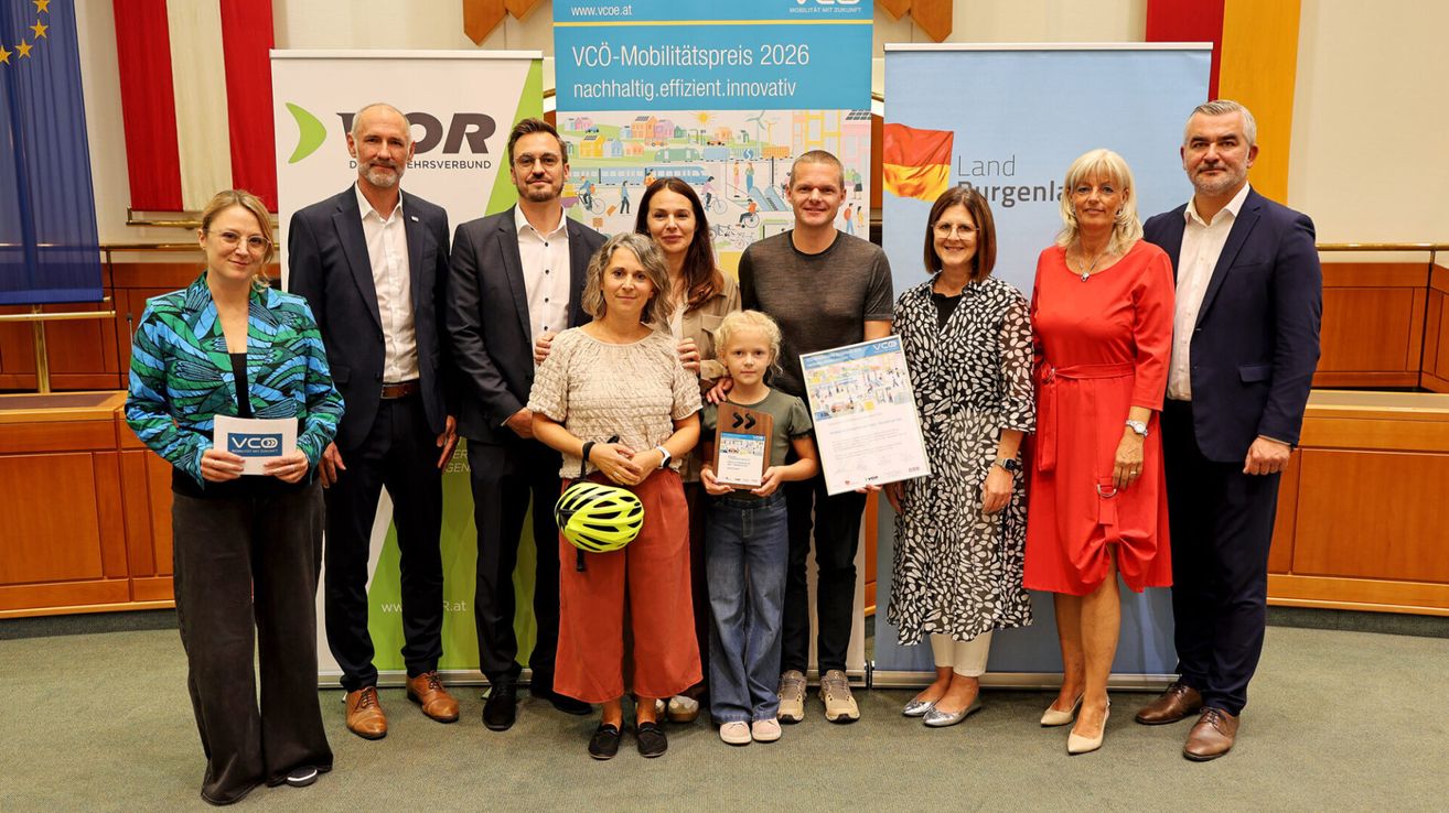 A group of people stand together in front of a backdrop with a banner. A young boy holds a certificate and a helmet. Behind him, a woman holds a certificate. On the left, a man holds a helmet. Behind them, two women and two men stand.
