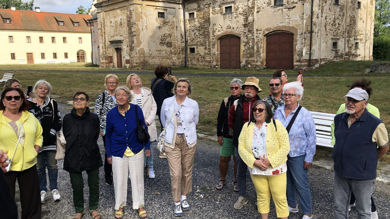 A group of elderly people are standing in front of an old building with stone walls and wooden doors. They are dressed in casual clothing and some are wearing glasses.