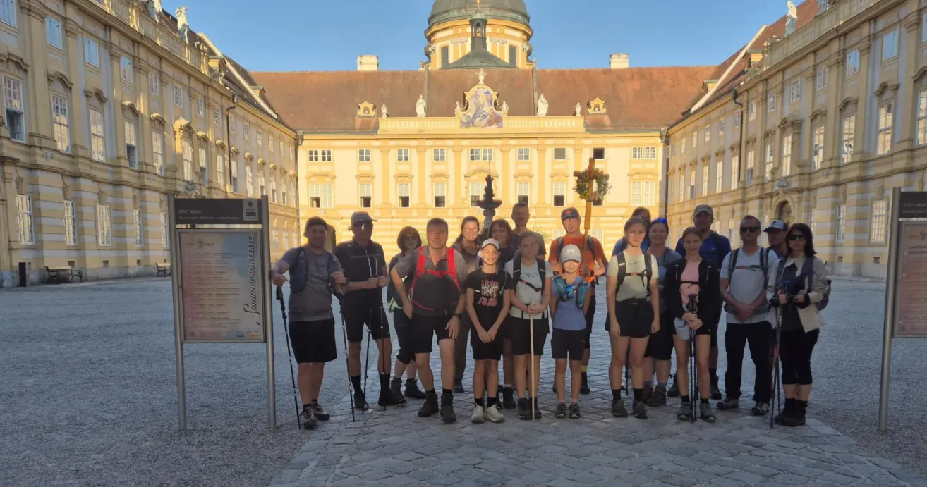 A group of hikers stands in front of a historic building with ornate architecture. They are all wearing hiking gear and some carry backpacks. The building has a dome-shaped roof and several windows.