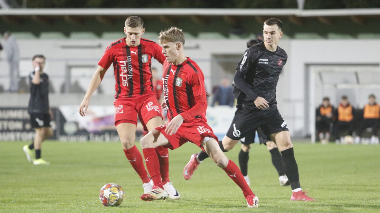 Drei Fußballspieler spielen auf einem Feld. Zwei tragen rote Trikots und einer schwarze. Sie jagen dem Ball hinterher.