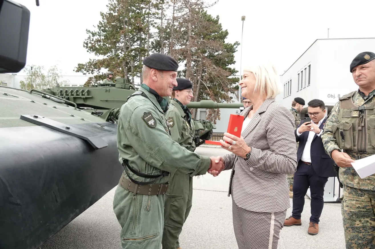 A military officer shakes hands with a woman in a suit in front of a tank. Behind them, a few people are standing, and a man is taking a picture.