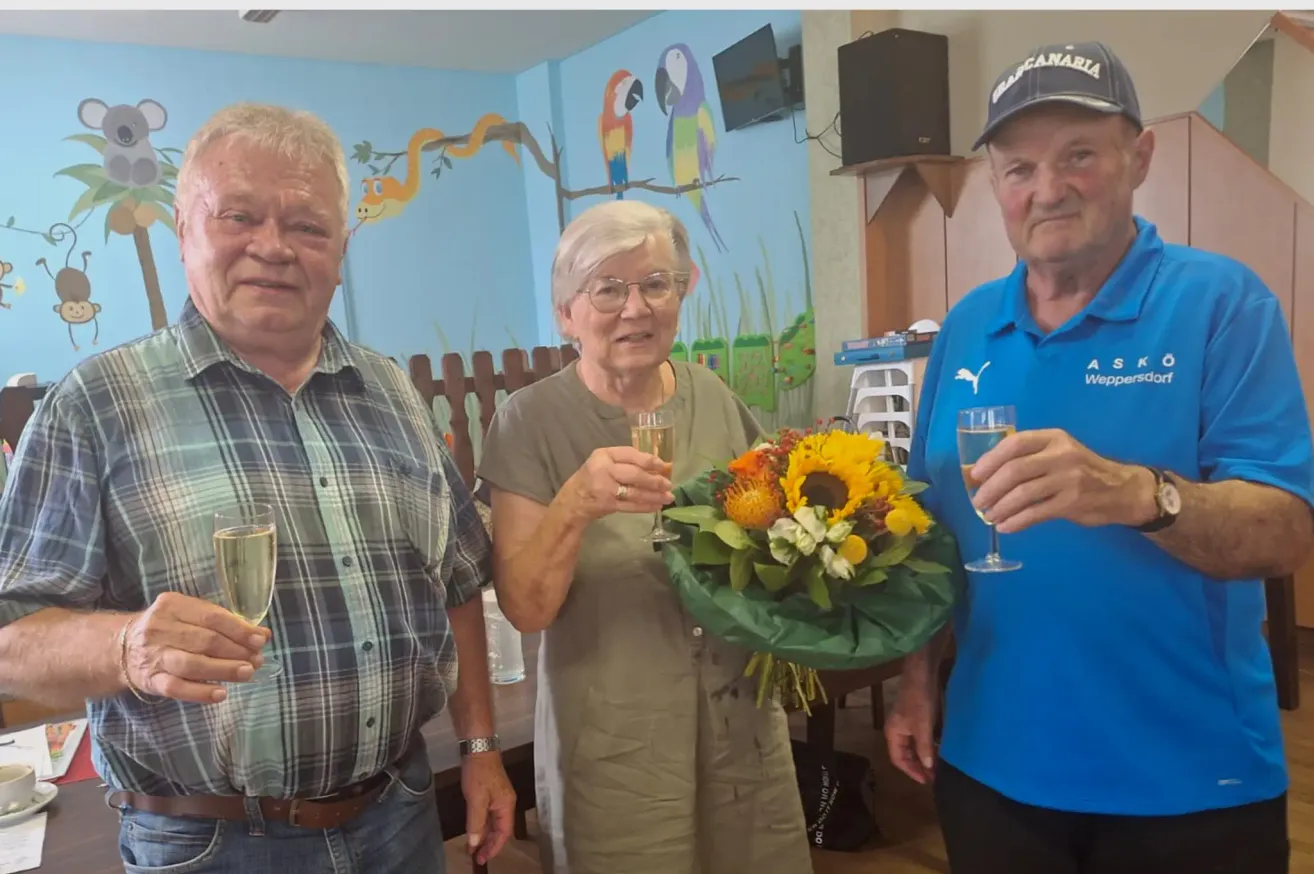Drei ältere Personen, zwei Männer und eine Frau, stehen zusammen, lächeln und halten Weingläser. Die Frau hält einen Blumenstrauß. Hinter ihnen befindet sich eine Wand mit einem Wandbild von Vögeln und Pflanzen.