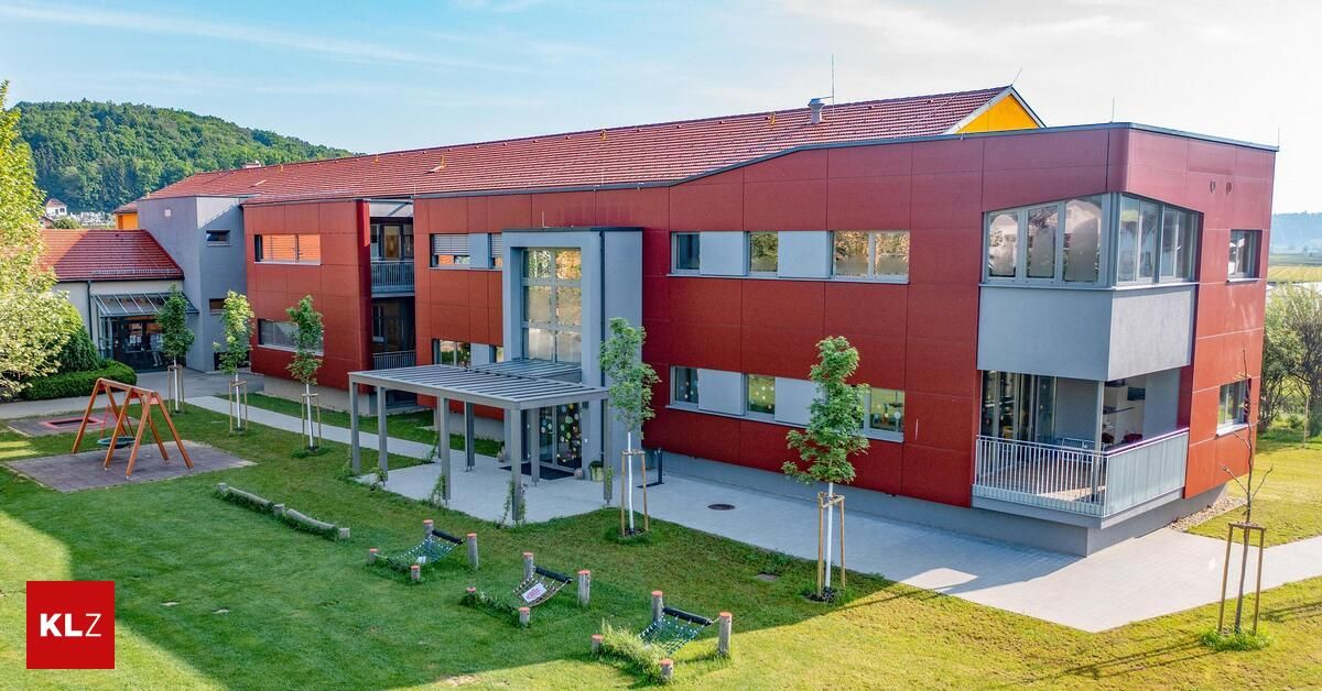 An aerial view of a modern red building with glass windows and a red roof. The building is surrounded by a green lawn with a few trees and plants. There is a walkway leading to the entrance of the building.