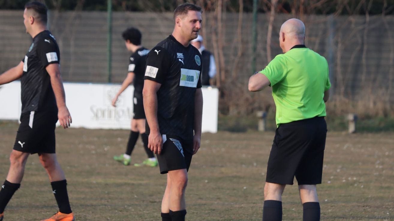 A soccer player in black uniform stands on a field, facing a referee in green. Behind them, another player walks in the background.
