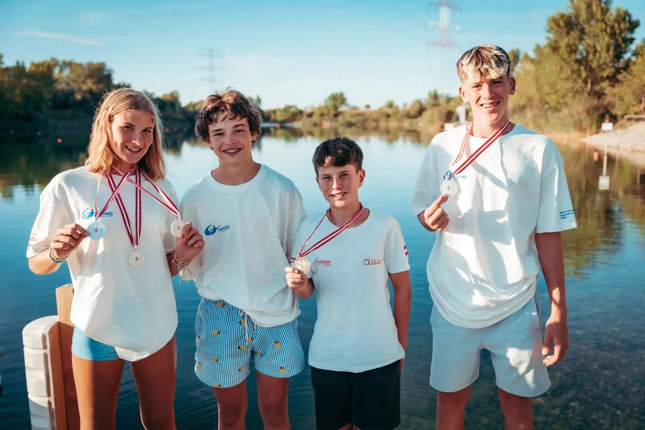 Four young individuals, likely athletes, stand by a lake, proudly displaying their medals. They wear matching white shirts and shorts, with two of them wearing blue shorts. The background features a serene lake and trees.