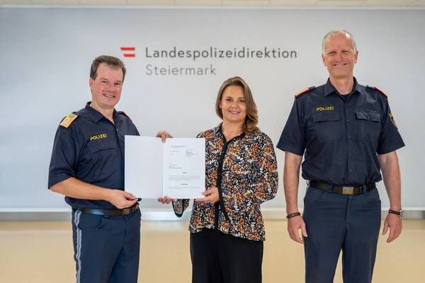 Three individuals in uniform stand in front of a wall with the text 'Landespolizeidirektion Steiermark'. A woman holds a document while two men stand beside her smiling.