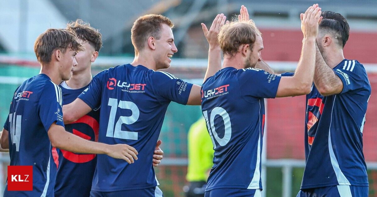 A group of soccer players in blue uniforms celebrate a goal. The player with the number 12 smiles and raises his hands.