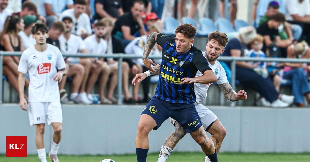 Two soccer players in action, one in a blue and black jersey with 'UMAFEX GRILL' and the other in white, both engaged in a tackle on a field.
