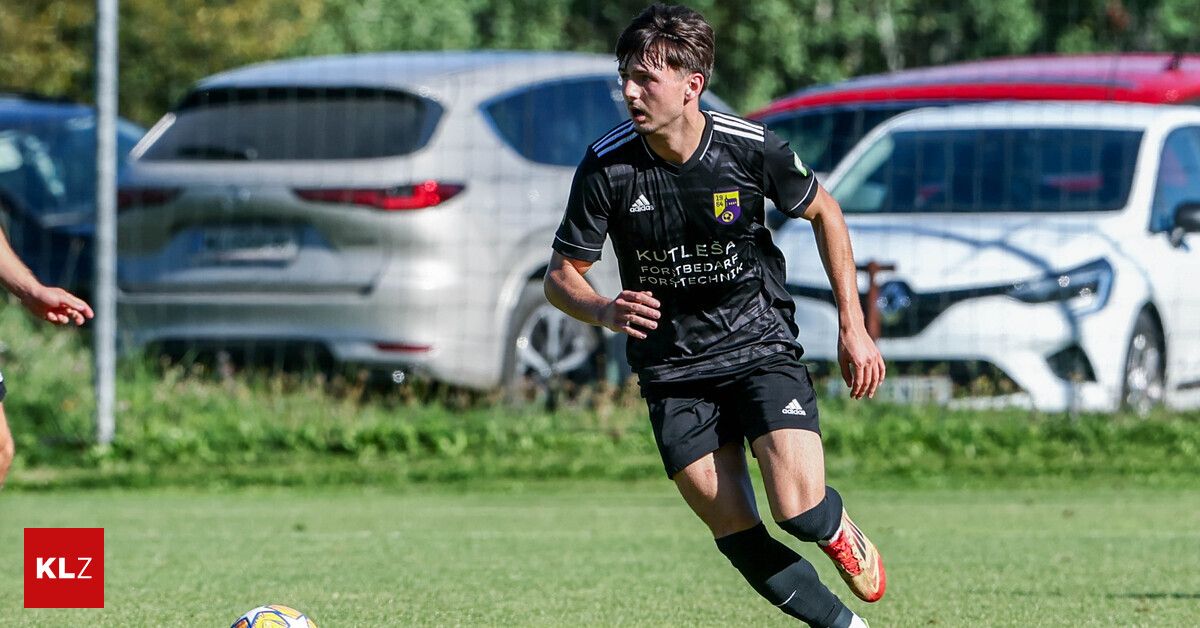 Ein Fußballspieler in schwarzer Uniform läuft auf dem Feld, mit einem Fußball in der Nähe. Im Hintergrund sind zwei geparkte Autos zu sehen.