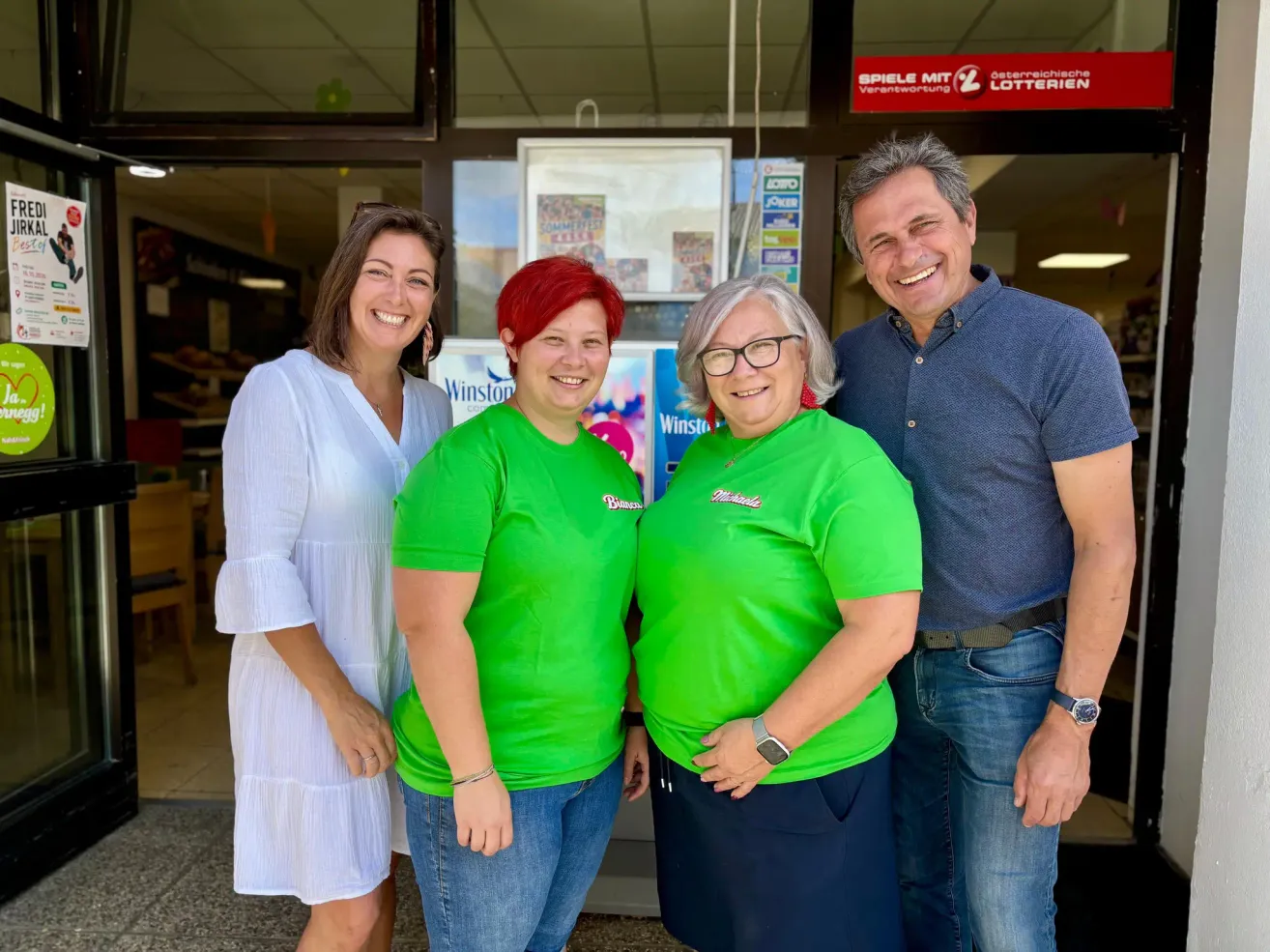 Vier Personen stehen vor einem Geschäft und lächeln für ein Foto. Zwei Frauen tragen grüne T-Shirts und zwei Männer tragen blaue und weiße Hemden. Hinter ihnen befindet sich ein Ladenschild und ein Plakat.