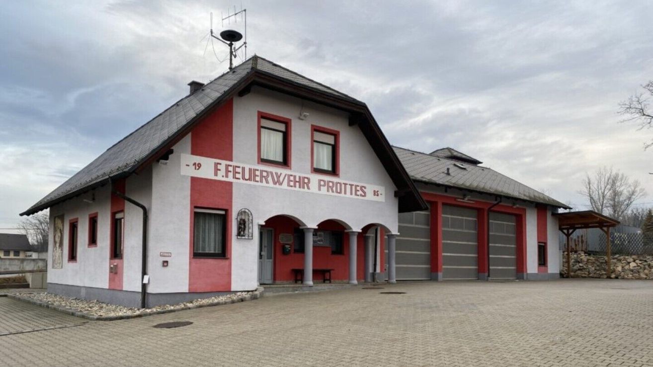 Ein rot-weißes Gebäude mit der Aufschrift F.Feuerwehr Prottes 85 an der Vorderseite. Es hat zwei große Garagen an der Seite und eine Bank davor.