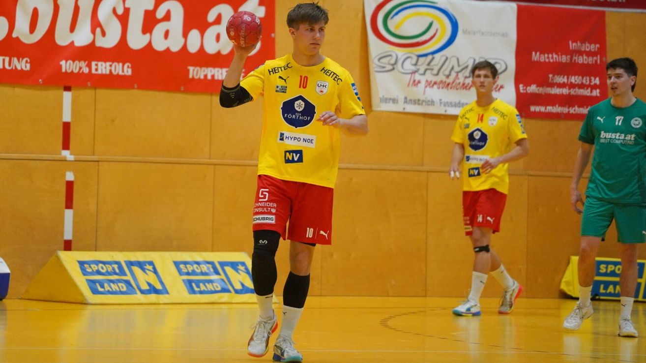 A male handball player in a yellow jersey with the number 10 is holding a red ball, preparing to throw it. He is standing on a court with a yellow floor.