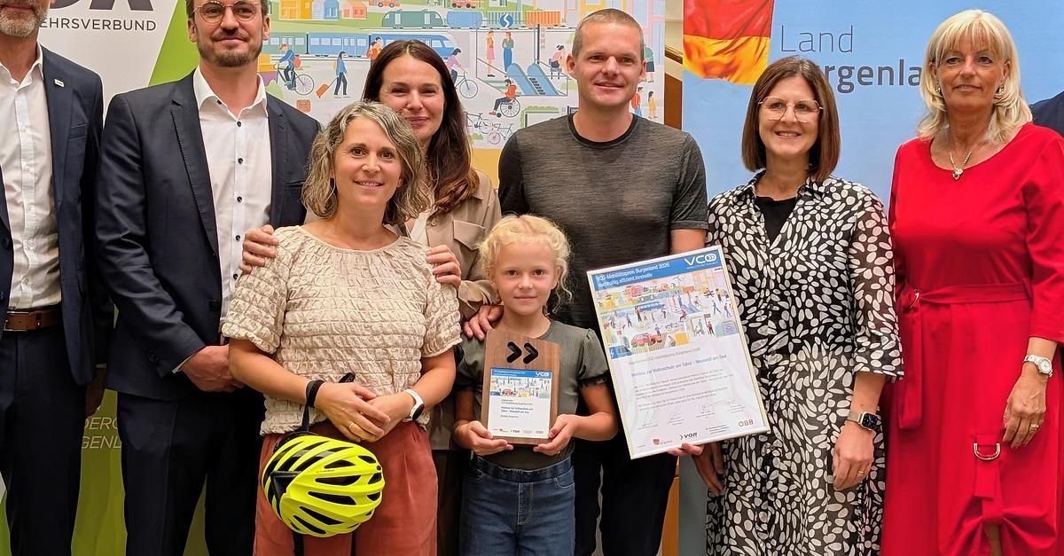 A group of people are standing together, smiling, and holding awards. A young girl in the center holds a certificate, while others hold certificates and a helmet. They are in front of a wall with a mural.
