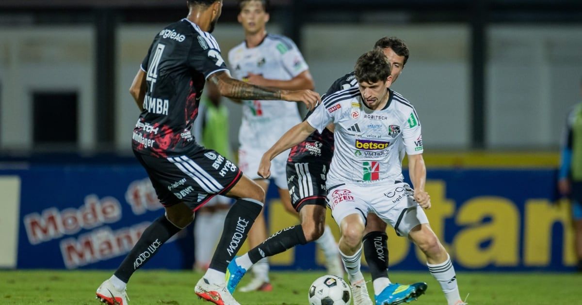 Drei Fußballspieler sind auf einem Feld, zwei in Weiß und einer in Schwarz. Der Spieler in Schwarz versucht, dem Spieler in Weiß den Ball zu stehlen. Der Spieler in Weiß schaut den Spieler in Schwarz an.