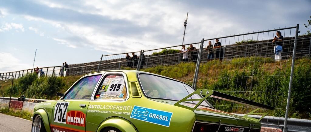 A green vintage car with the number 752 on its side is parked on a hill with a metal fence. The car has a sticker with the text 'AUTOLACKIEREI' and 'GRASSL'. There are people standing on the side of the hill.