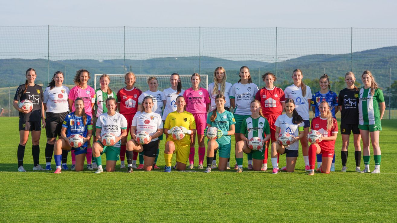 A group of female soccer players in various team jerseys pose for a photo on a field with a fence and mountains in the background.