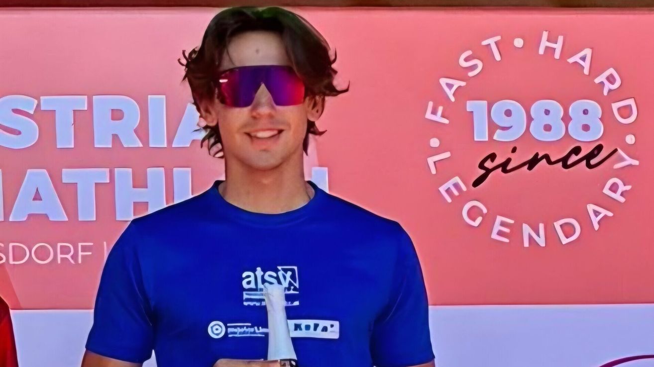 A man with brown hair wearing sunglasses and a blue t-shirt with white text is smiling and holding a bottle. Behind him is a pink wall with white text.