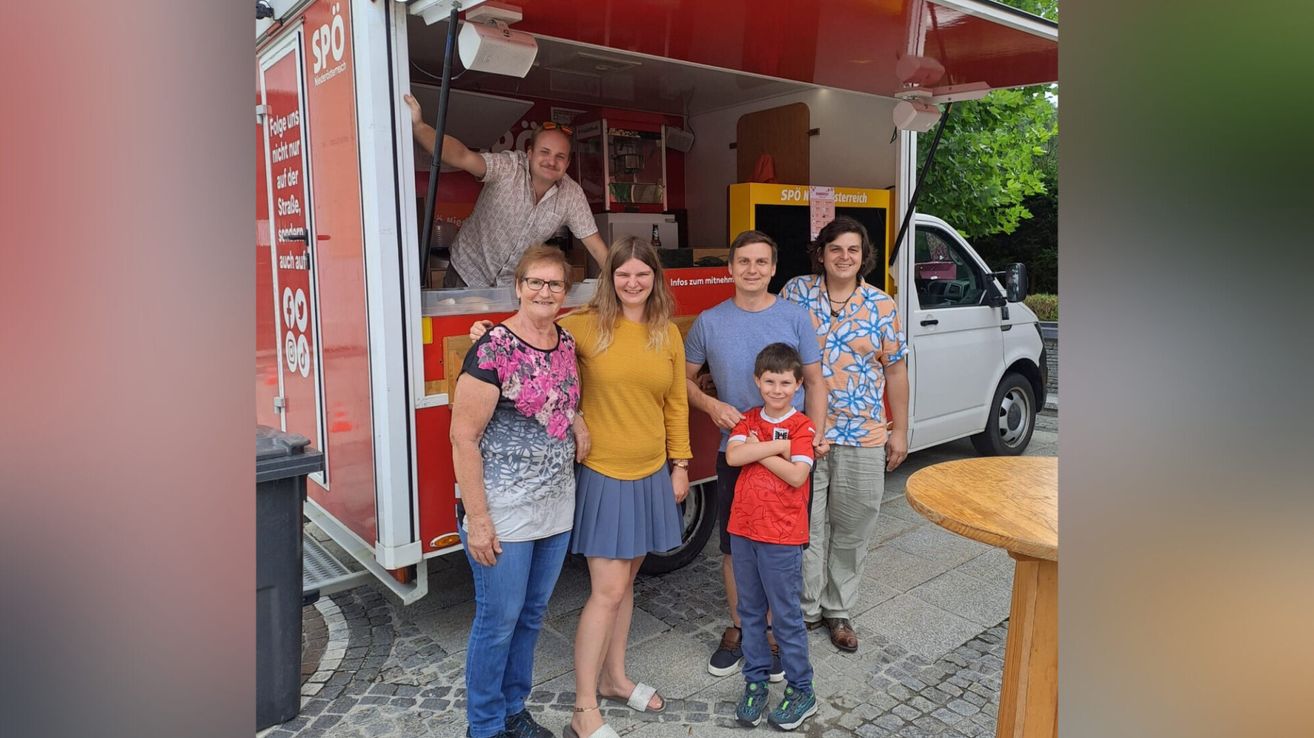 A family stands in front of a food truck, with two women, two men, and a young boy smiling. The man inside the truck wears sunglasses on his head.