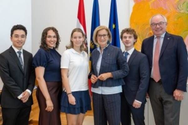 Eine Gruppe von Menschen, darunter eine Frau und vier junge Personen, steht gemeinsam vor einer Wand mit Flaggen und einem Gemälde.