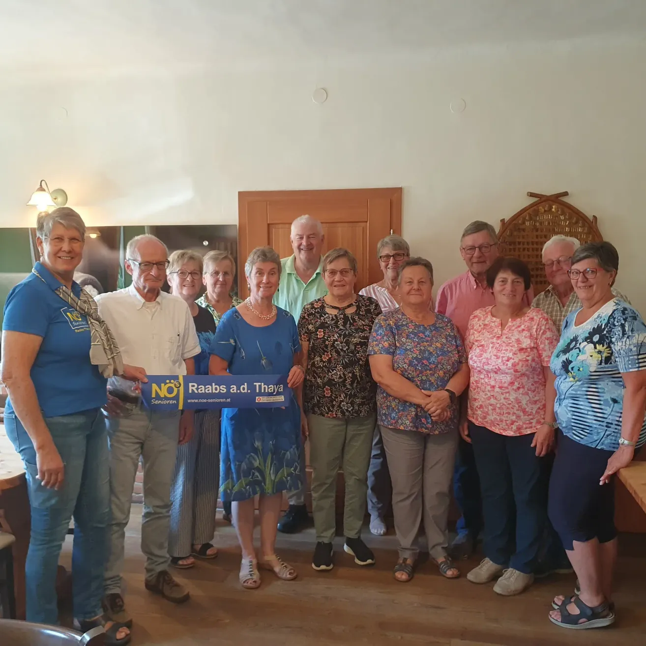 A group of people stand together in a room, smiling and posing for a photo. A man holds a blue sign with white text. The room has wooden floors and white walls.