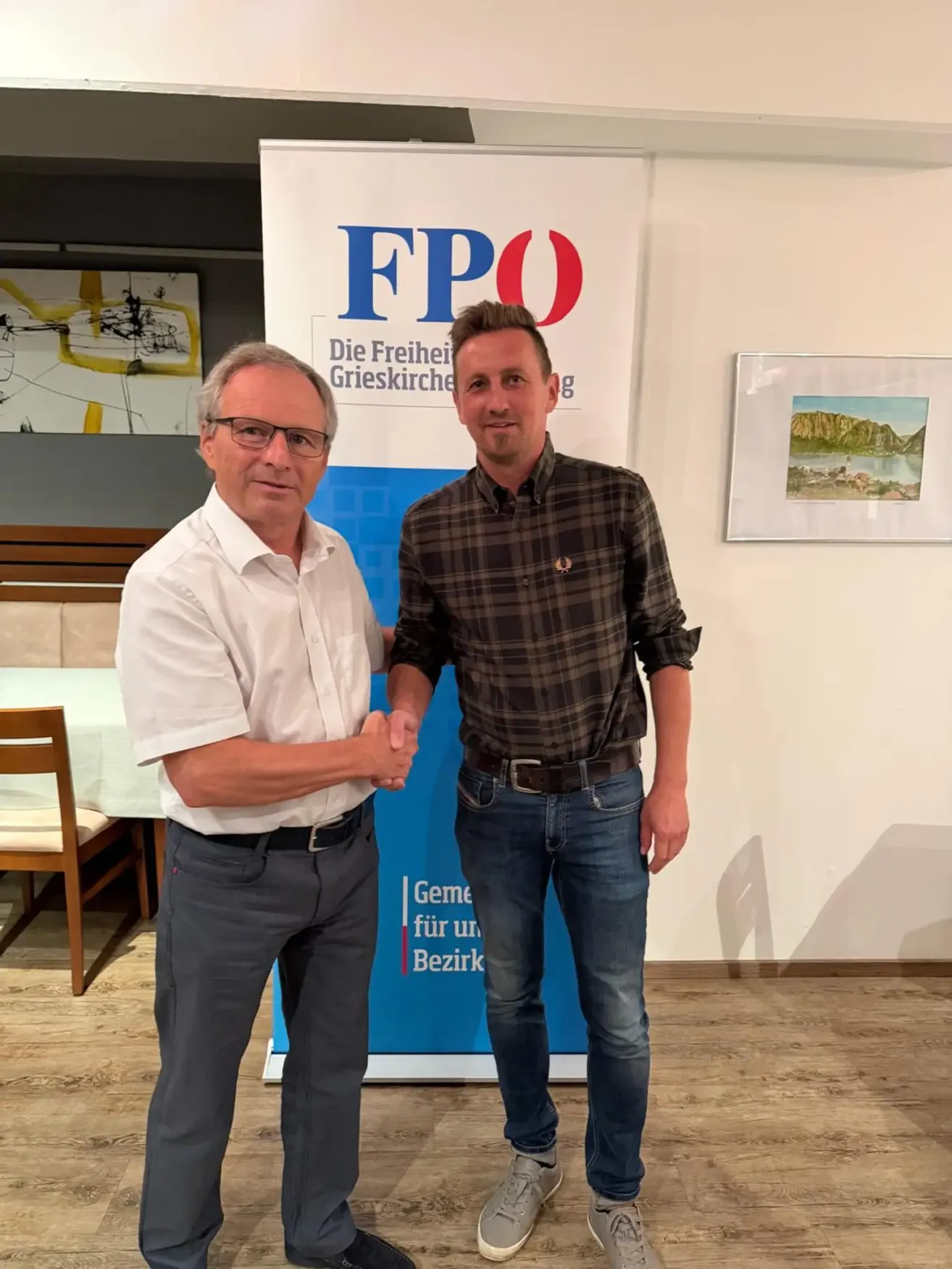 Zwei Männer stehen nebeneinander und schütteln sich die Hände vor einem Banner, auf dem 'FPO' steht. Der Mann auf der linken Seite trägt eine Brille und ein weißes Hemd, während der Mann auf der rechten Seite ein kariertes Hemd und Jeans trägt. Hinter ihnen ist eine Wand mit einem Bilderrahmen und einem Tisch mit einem Stuhl.