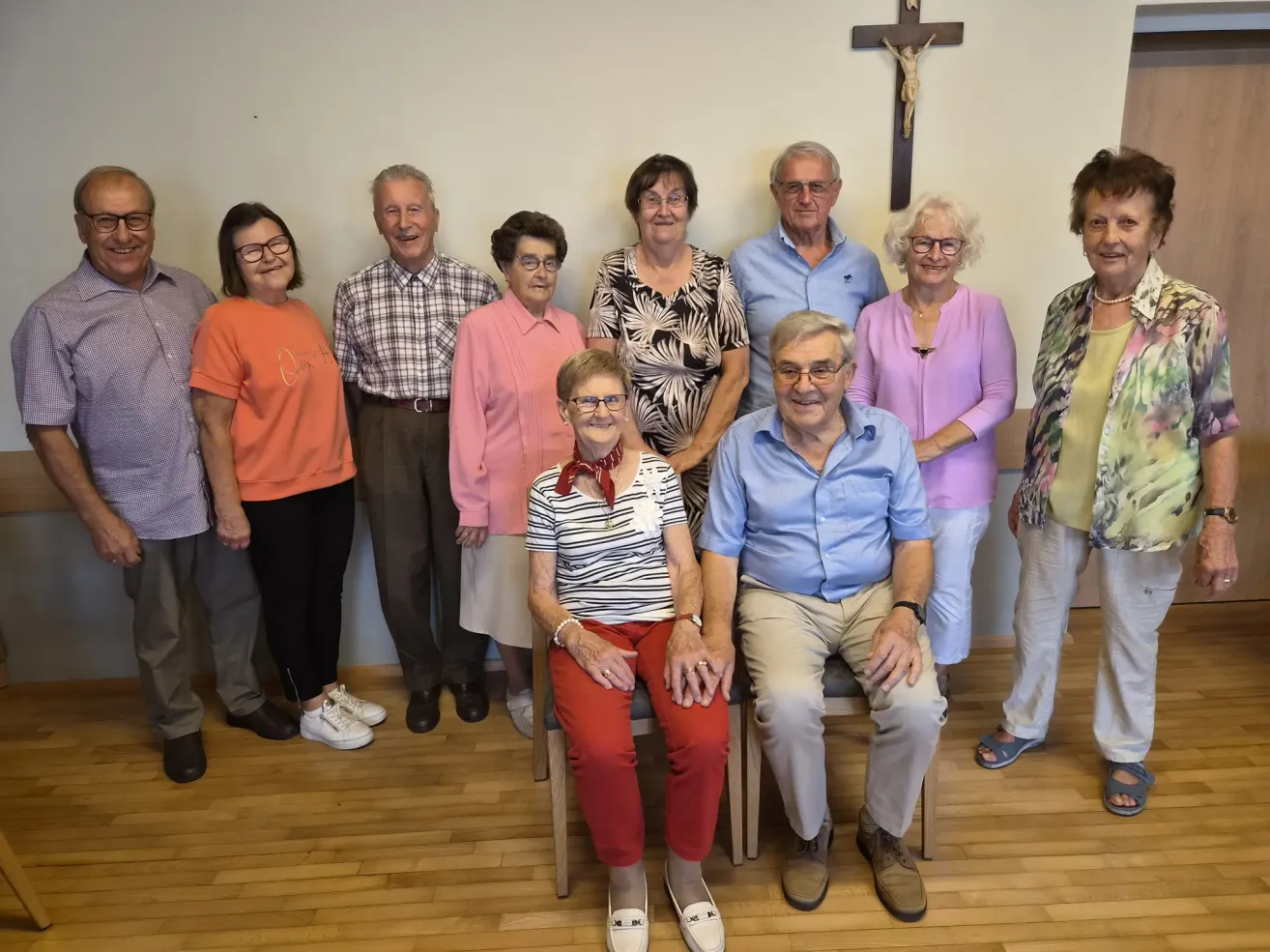 A group of elderly people, both men and women, are posing for a photo in a room with a cross on the wall.