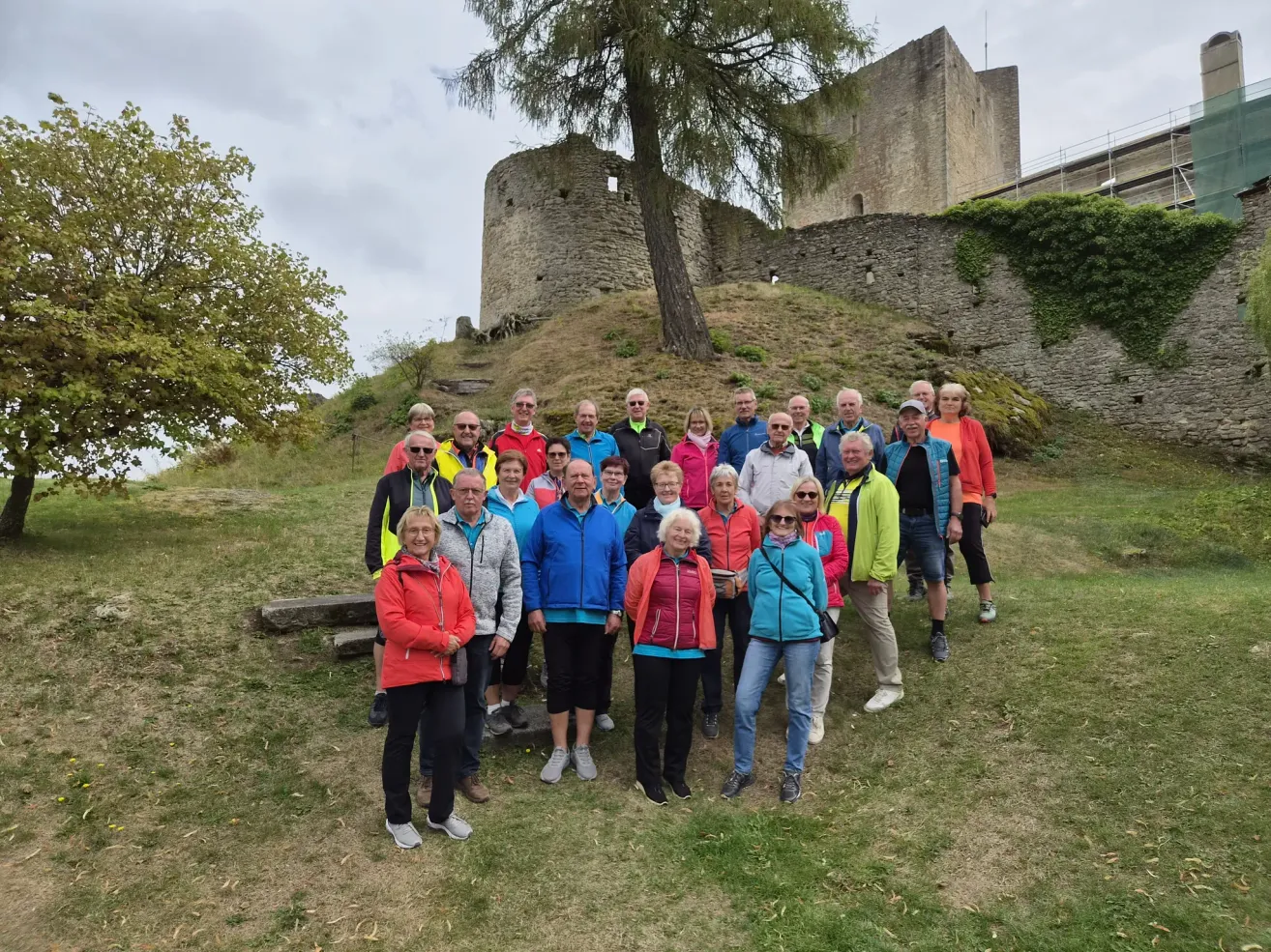 A group of people dressed in various colors pose for a photo in front of an old castle on a hill.