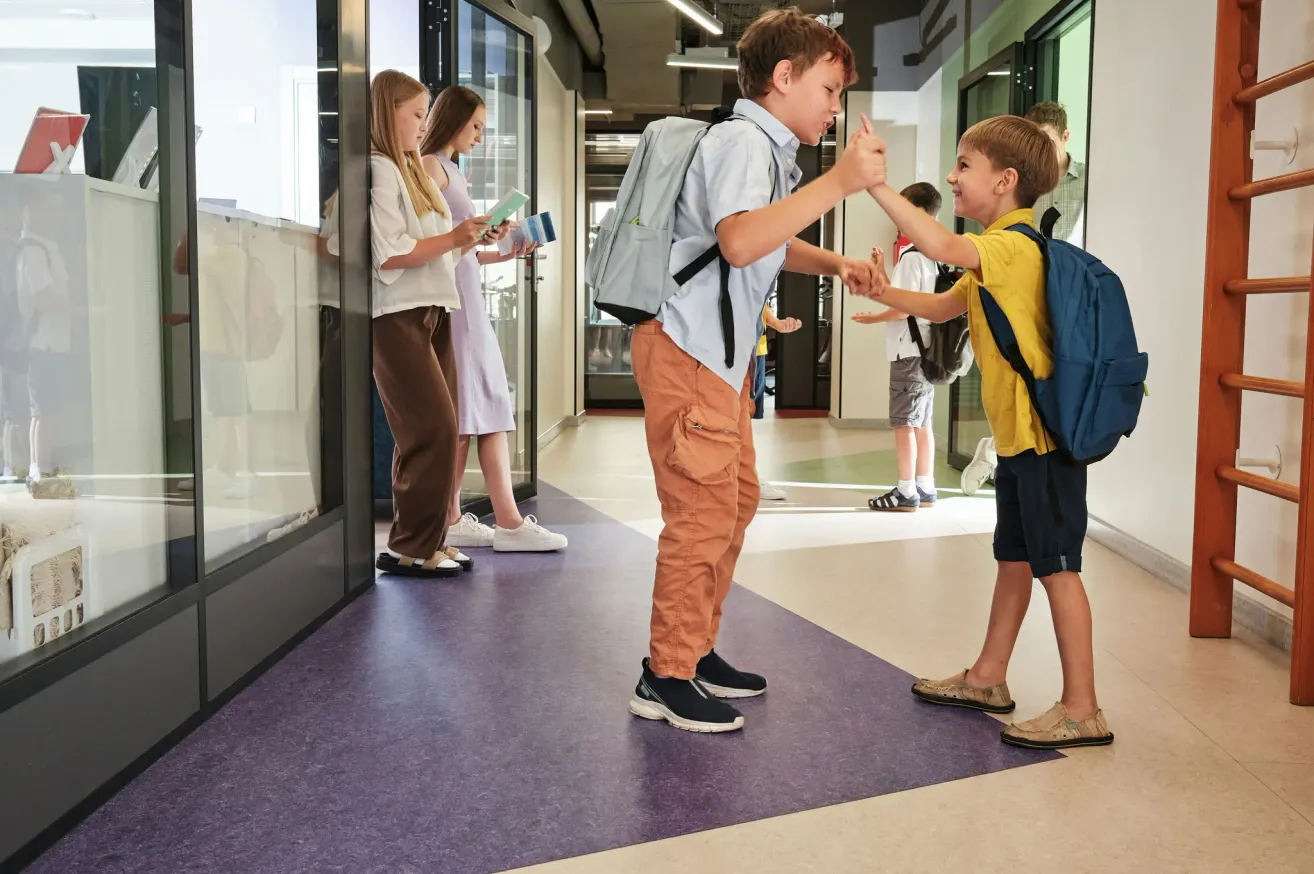 Zwei Jungen mit Rucksäcken geben sich gegenseitig einen High-Five in einem Schulflur. Hinter ihnen stehen zwei Mädchen in der Nähe einer Glastür, eines hält ein Buch. Andere Schüler sind im Hintergrund.