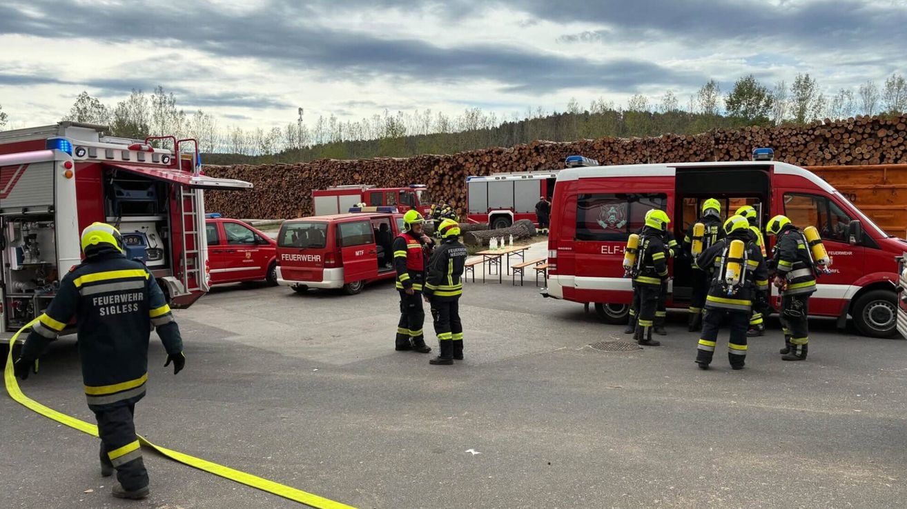Firefighters in red uniforms stand in a parking lot, with fire trucks and benches in the background.