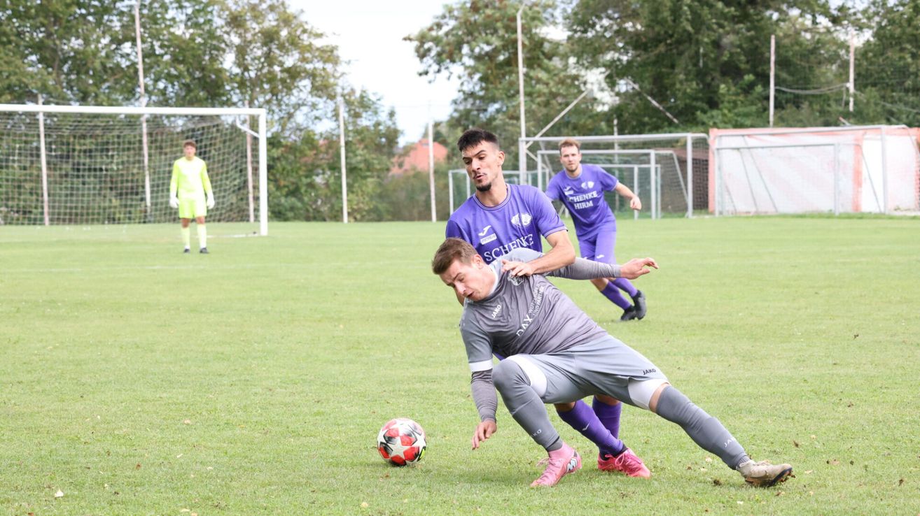Zwei Fußballspieler in lila und grauen Trikots sind auf einem Fußballfeld im Clinch. Der Spieler in Lila versucht, dem Spieler in Grau den Ball zu stehlen.