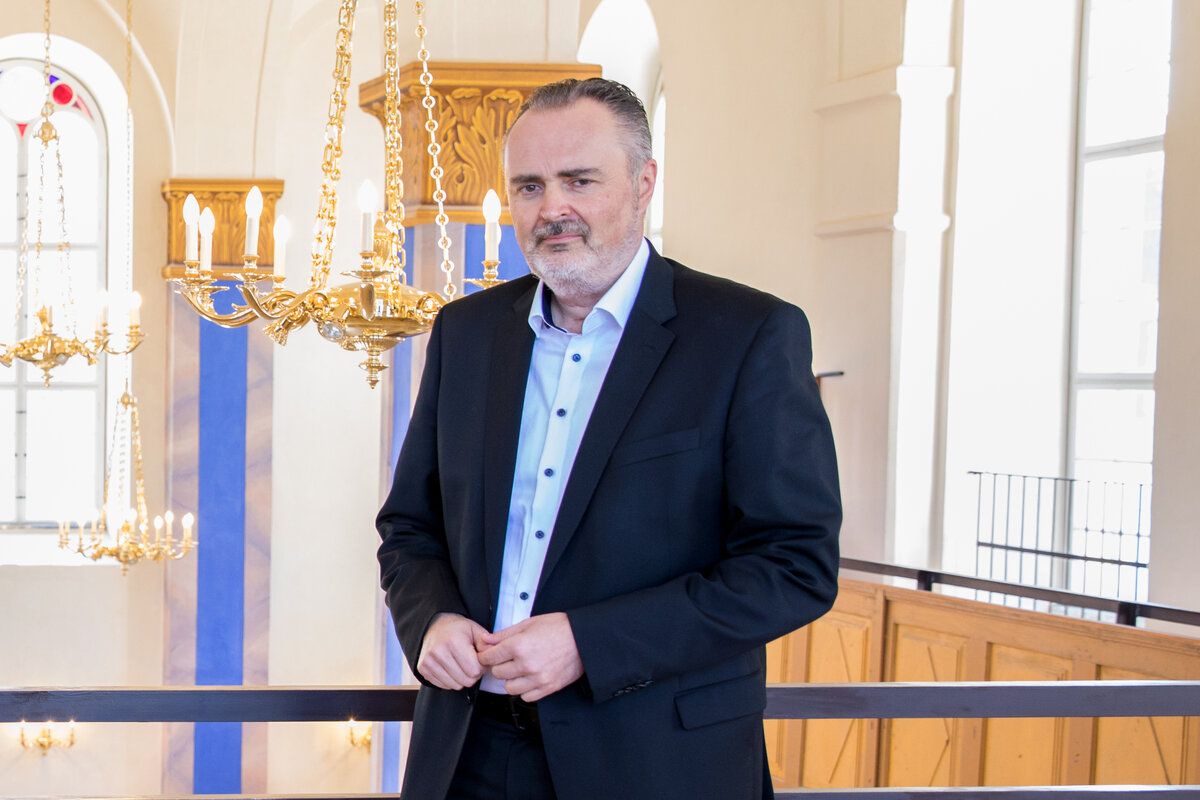 Ein Mann in einem schwarzen Anzug steht in einer Kirche und sieht zur Seite. Hinter ihm hängt eine goldene Kronleuchter an der Decke.
