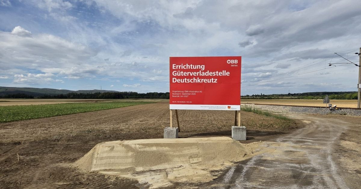 A red sign in a field reads 'Errichtung Güterverkehrsstelle Deutschkreuz'. The sign is on a concrete base with a dirt mound in front. The sky is cloudy.