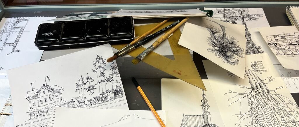 A workspace with various sketches and drawing tools. A black palette with watercolors, brushes, and papers with drawings of trees, buildings, and a tower.