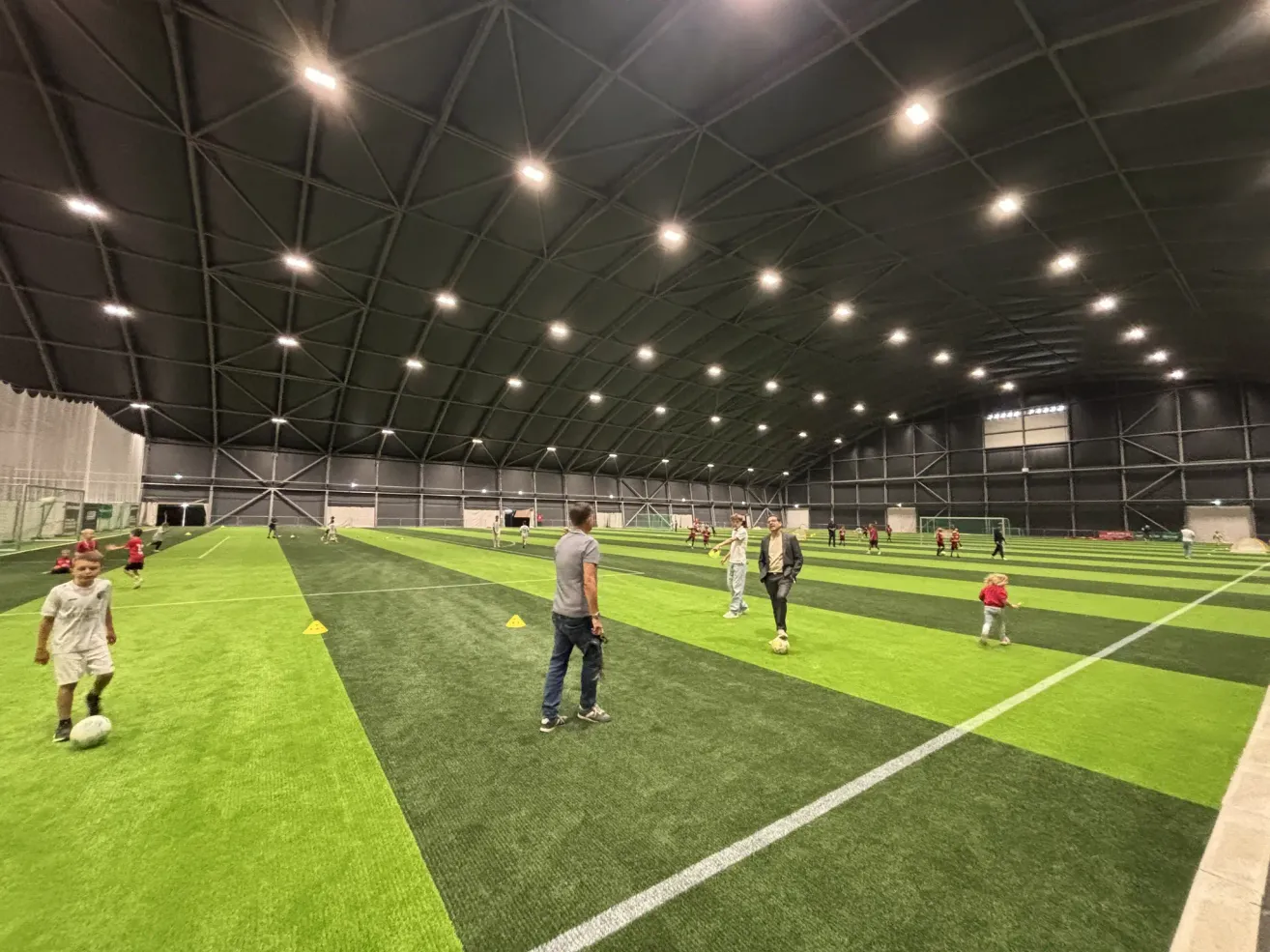 Eine Gruppe von Menschen spielt Fußball auf einem überdachten Feld mit grünem Rasen und weißen Markierungen. Das Feld ist von einer hohen Decke mit Lichtern umgeben.