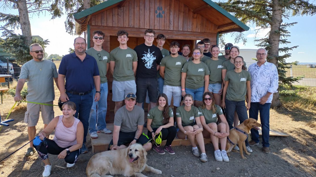 Eine Gruppe von Menschen in grünen T-Shirts, einige mit Brille, posiert für ein Foto vor einer Holzhütte. Ein Golden Retriever liegt in der Nähe auf dem Boden.