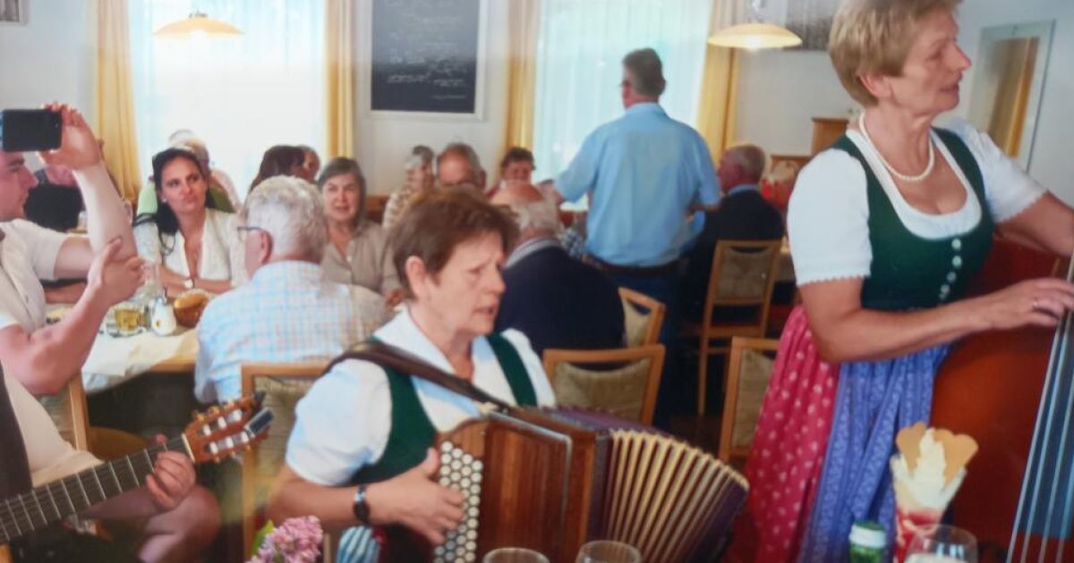 Eine Frau spielt Akkordeon in einem überfüllten Restaurant, mit mehreren sitzenden und stehenden Personen um sie herum.